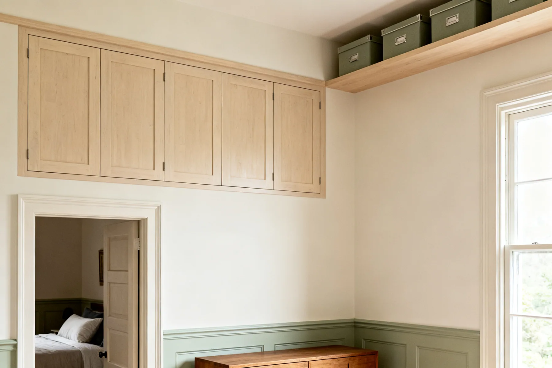 The band above 1.9m that furniture never reaches A bedroom with closed cabinets fitted above the doorway continuing as a high shelf around the corner holding lidded storage boxes
