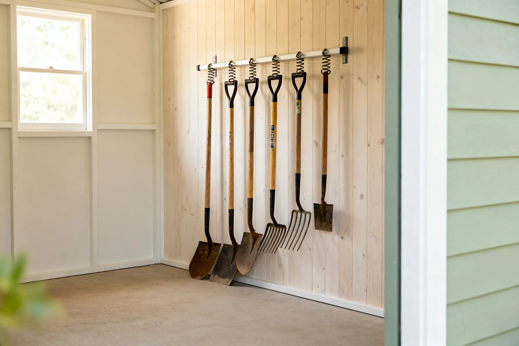 Long handles belong on a rail A shed wall with long-handled garden tools hung upright on a rail with spring grips, floor beneath clear