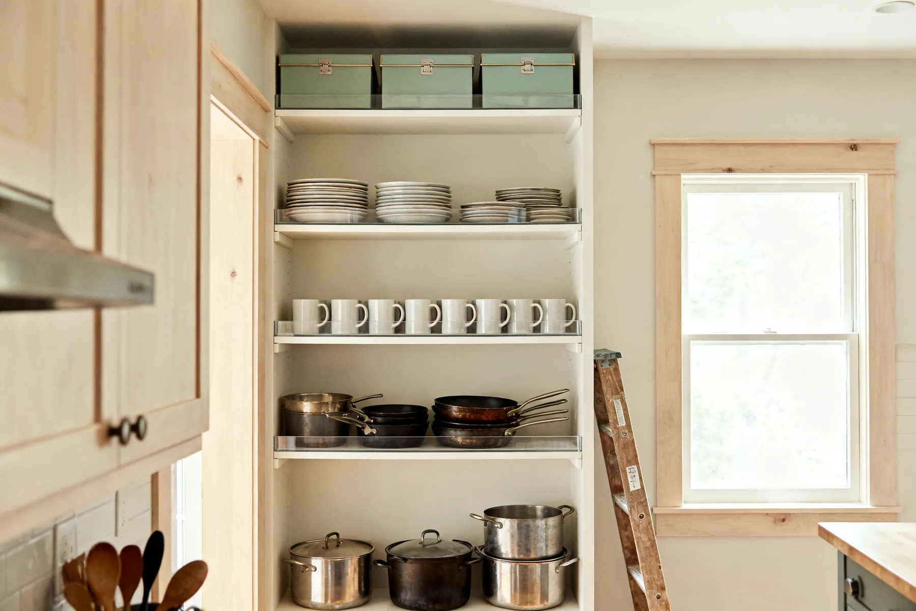 Zoned by how often things get used A tall kitchen storage wall loaded in three bands: everyday plates at chest height, heavy pans low down, and lidded boxes on the top shelf with a step stool nearby