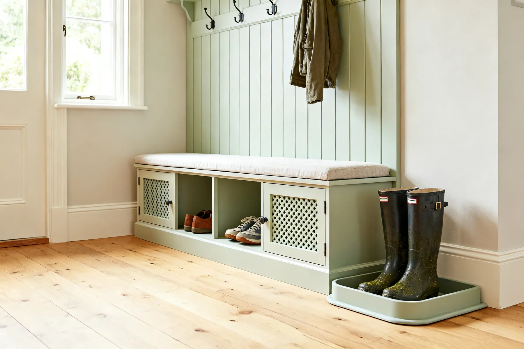 A bench, because nobody puts boots on standing up A hallway bench with a padded seat and open ventilated shoe cubbies underneath each holding one pair, coat hooks above, and a raised-lip boot tray with wellingtons beside it